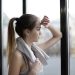 Side view of thoughtful young female athlete in sportswear with towel around neck standing near window with elbow on glass and looking away pensively while resting after training in modern gym